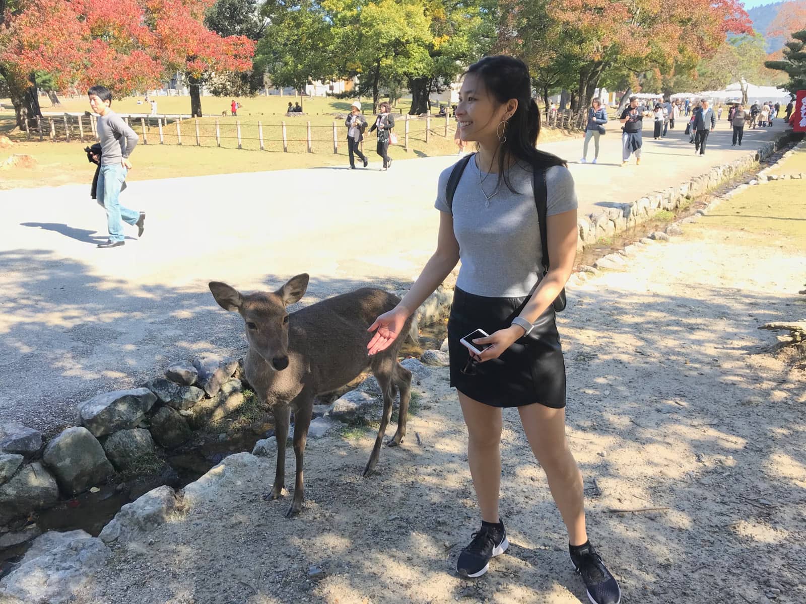 A woman in a grey t-shirt, black skirt and running shoes, standing next to a deer, obviously posing and smiling but not looking at the camera