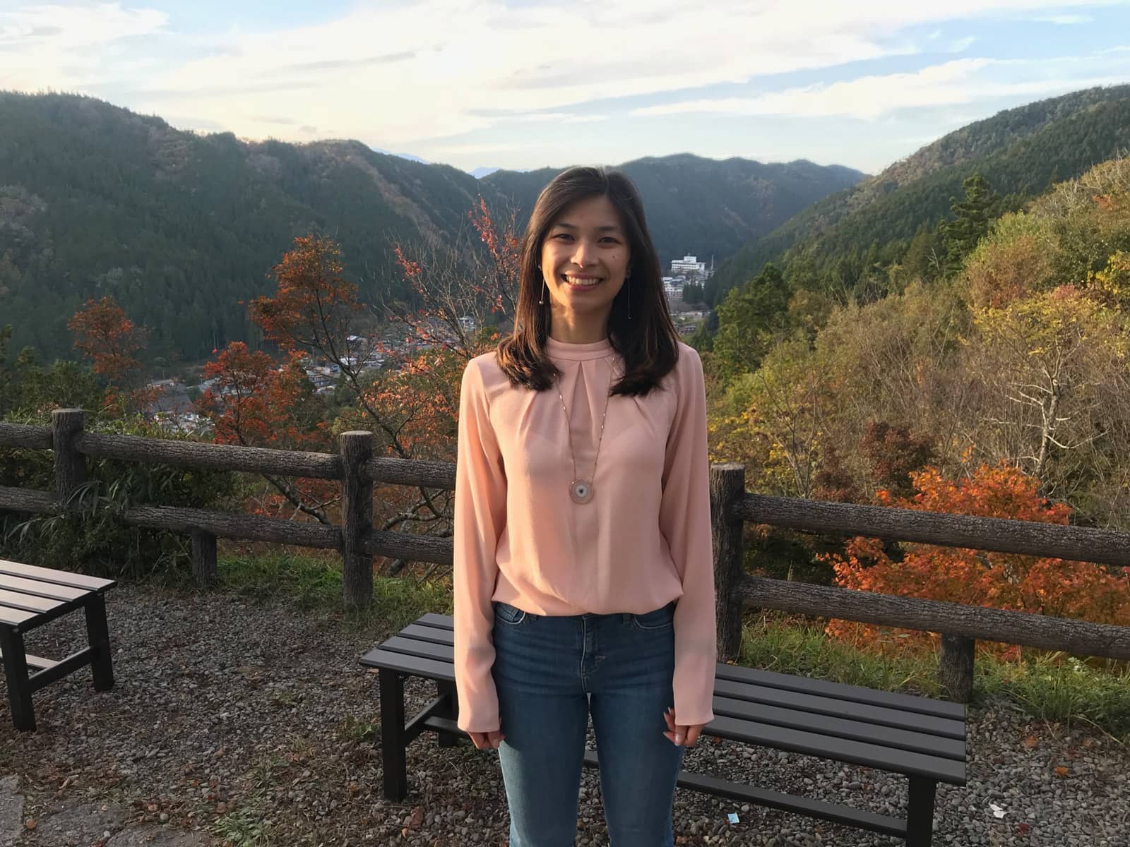 a woman in a long sleeved pink blouse and blue jeans, at a lookout with a remote town and autumn leaves in the surroundings