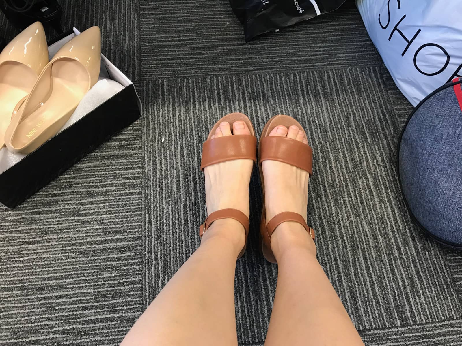 A point-of-view photo of a woman’s feet with brown sandals on. In the background is a pair of nude-coloured heels