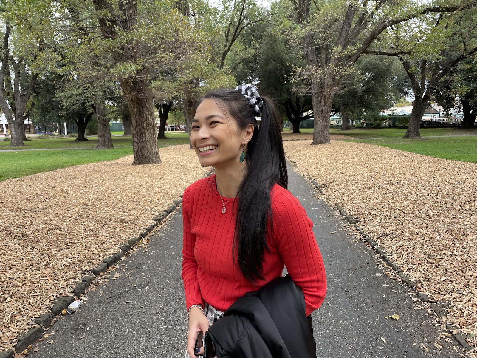 An Asian woman standing in a park, grinning. She has her long dark hair tied in a ponytail and a black-and-white gingham scrunchie, and is wearing a long sleeved red top and carrying a black jacket.