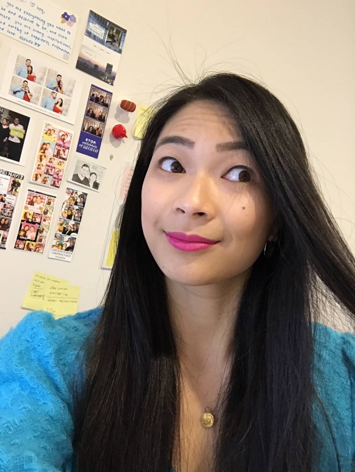 A woman with long dark hair, smiling, taking a selfie. She’s wearing a blue top and there is a wall behind her with some photos