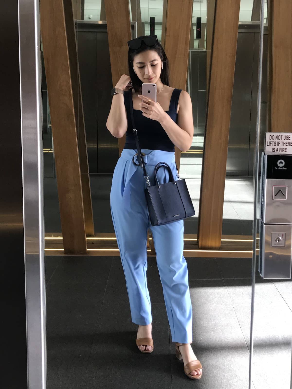 A woman taking a selfie in a mirror. She’s wearing a navy sleeveless top with sky blue tapered pants.