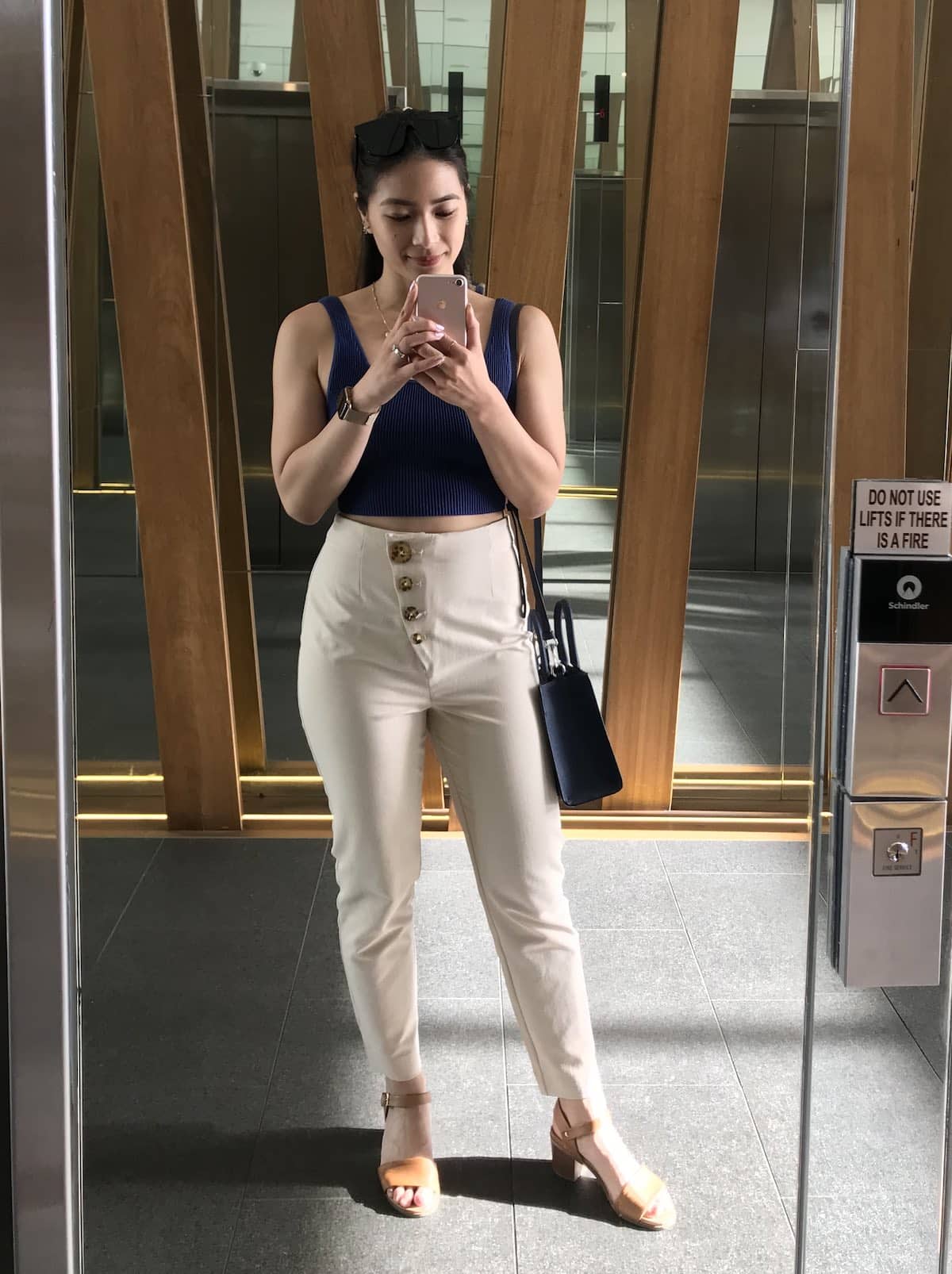 A woman wearing a navy top and beige pants, taking a selfie in a mirror. She has dark sunglasses on top of her head and is also wearing tan sandals.