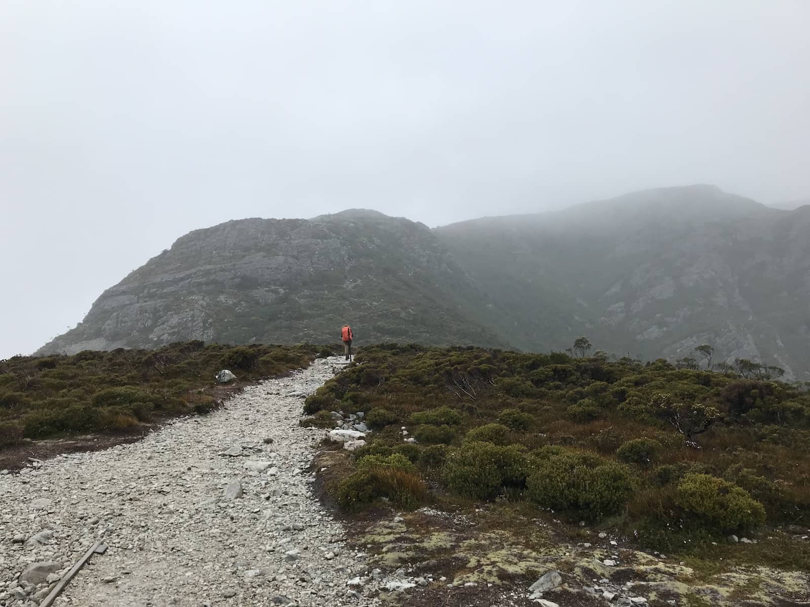 A gravel path on a hike, leading up to a mountain. A hiker with a bright backpack can be seen up ahead.