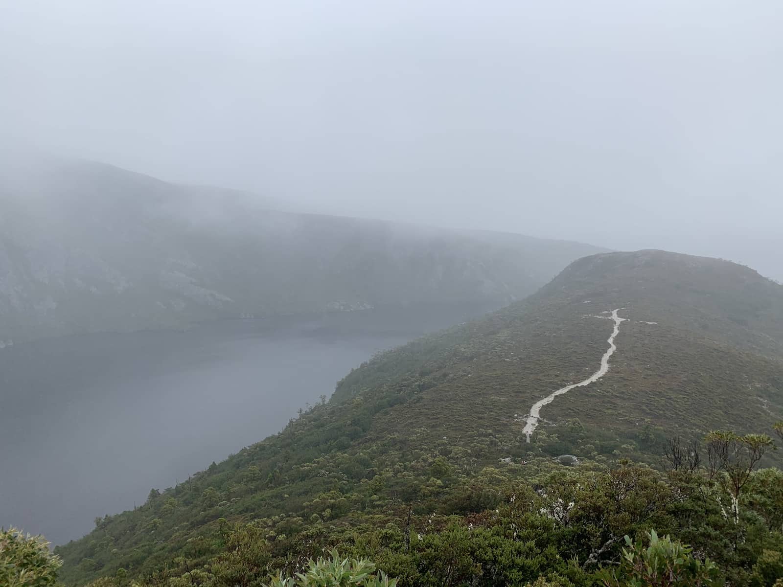 A view downwards from a mountain, showing a thin trail leading into the distance. It is a very cloudy day