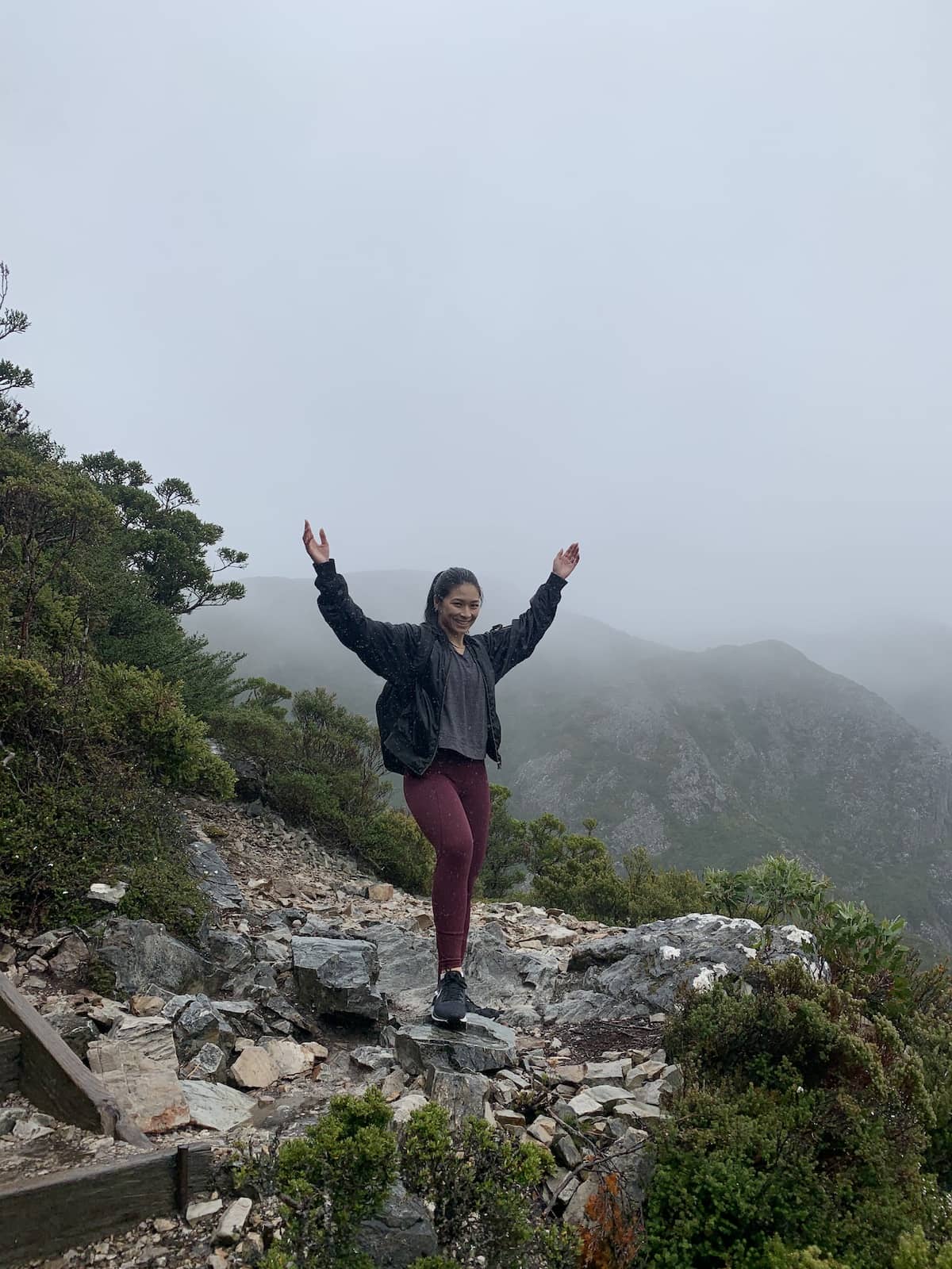 A woman with dark red leggings and a black jacket, with her hands in the air. She’s standing on mountainous terrain and it’s really cloudy in the background