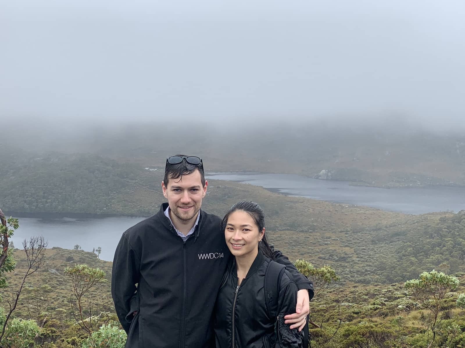 A man and woman with their arms around each other, with some lakes in the background. They are damp from the rain