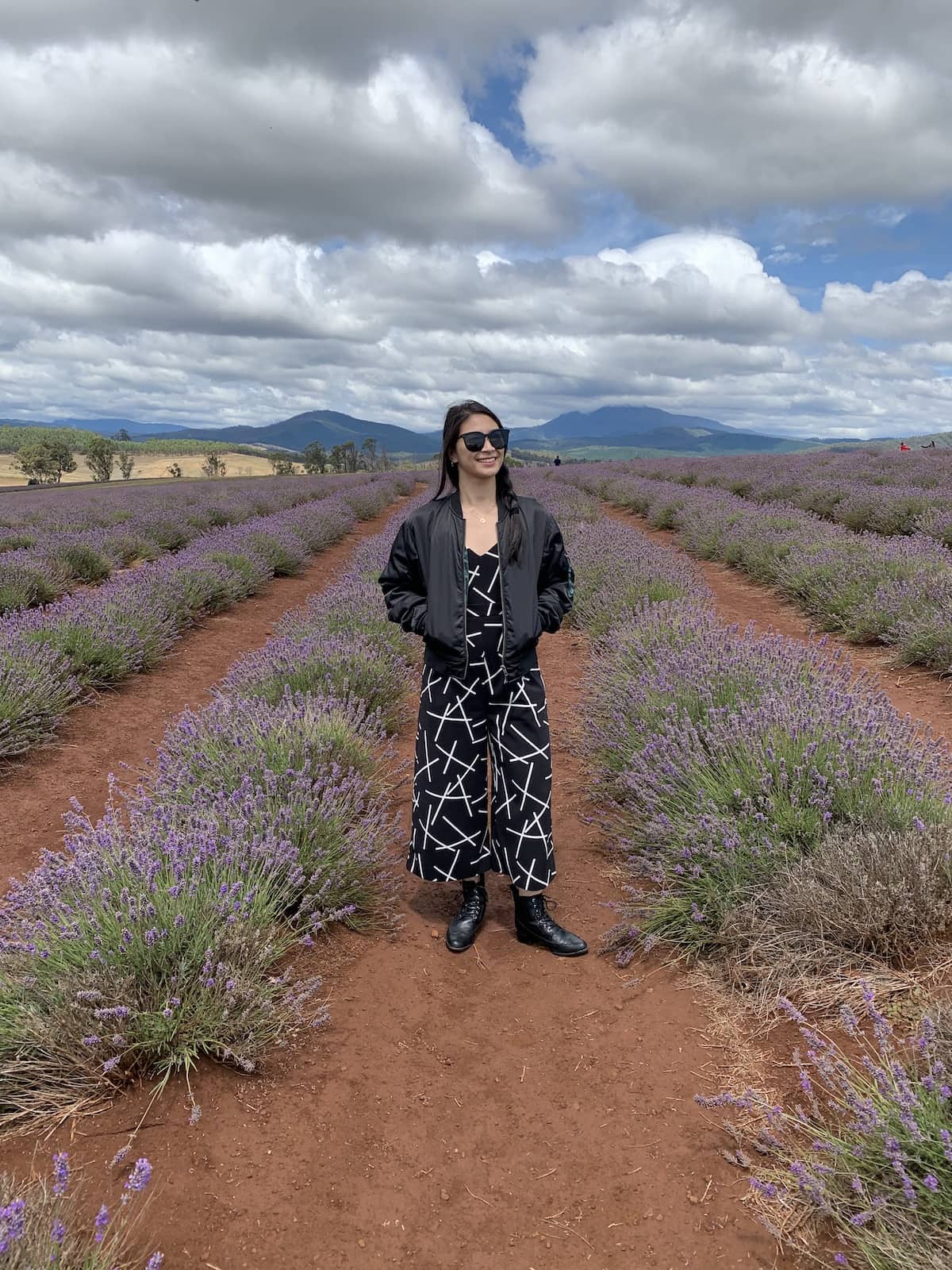 A woman in a black patterned jumpsuit and black bomber jacket, standing between rows of planted lavender. She is wearing black sunglasses.