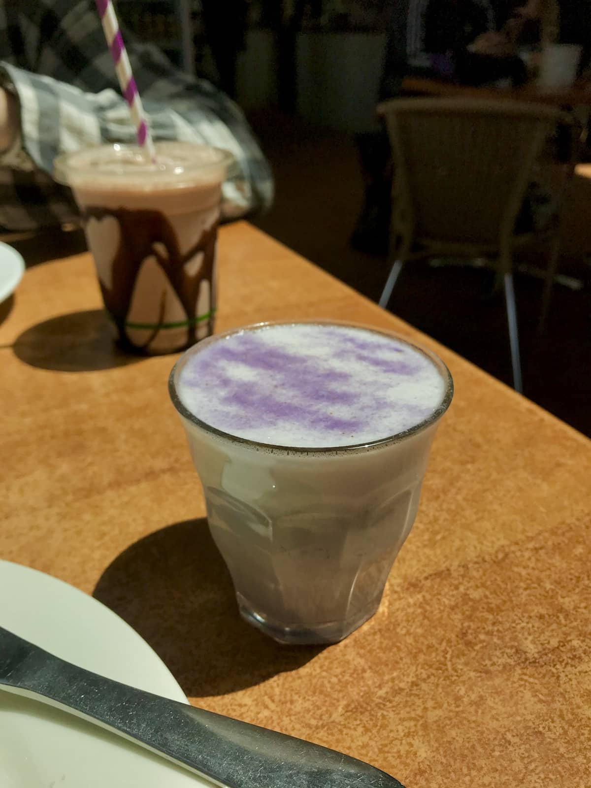 A glass cup with a milky drink of a light purple colour, sitting on a table in the afternoon sun