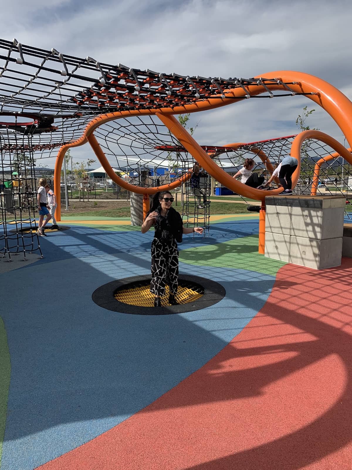 A woman in a black patterned jumpsuit, wearing sunglasses, jumping in a shallow hole in a children’s playground. The hole is covered with flexible structure that makes the surface look bouncy