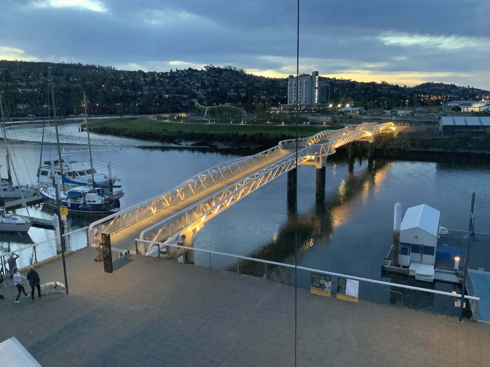 A short pedestrian bridge going over a river, to another small island with a large playground and a hotel resembling a silo. It’s evening and the bridge is lit up.