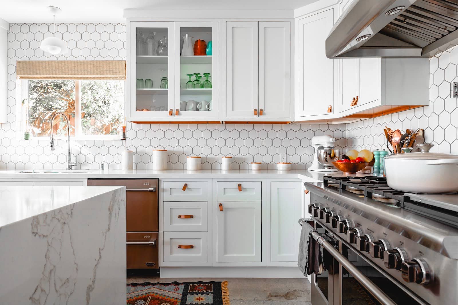 A brightly-lit kitchen with white walls of honeycomb-shaped tiles. The kitchentop on the left is made of white marble, and an oven sits on the right. Ahead, some white cabinets can be seen, some with windows