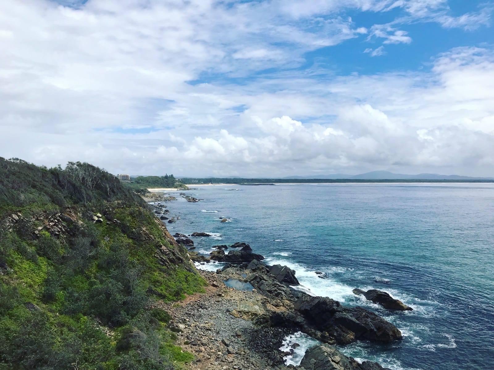 A view from a high vantage point of a high part of grassy bushland leading to the ocean. A rock beach separates the land from the water. The sky is blue but has many clouds