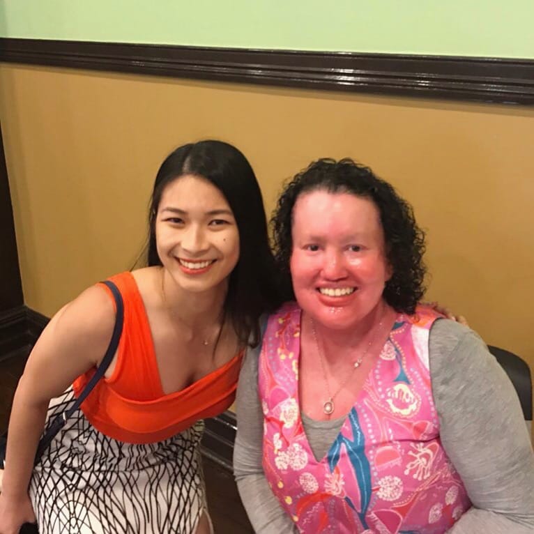 Two women smiling, one has light skin and dark straight hair, and the other has dark curly hair and pink skin. They are both wearing bright orange and pink.