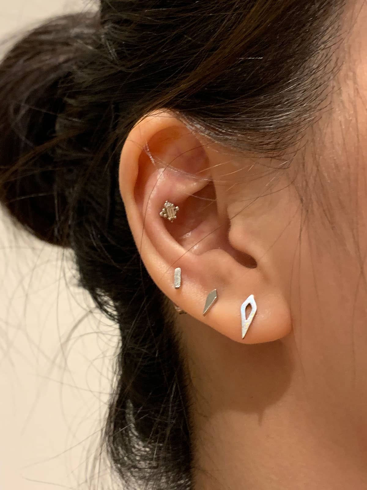 A closeup of a woman’s ear. She has dark hair that is tied back. Her ears have three silver earrings on the lobe and a smaller silver one near the inner ear.