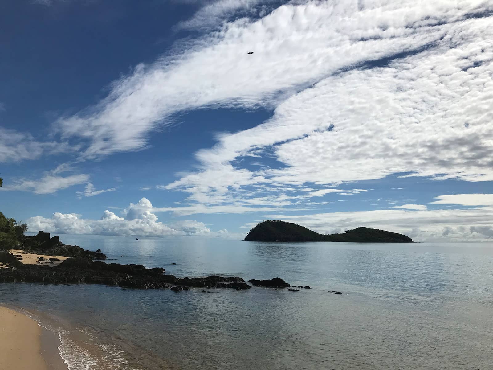 A view of the shore of a beach, with vast stretches of white clouds in the blue sky. The water by the beach is blue and there are some rocks in the shallow part of the water.