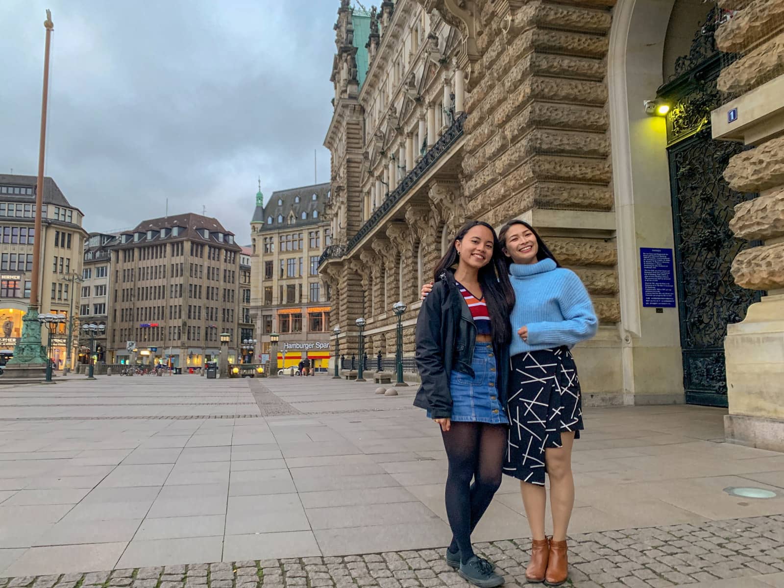 Two women with dark hair with their arms around each other, smiling. One is dressed in a denim skirt and black jacket, the other in a blue sweater and black patterned skirt. They are in a quiet and cloudy town square.