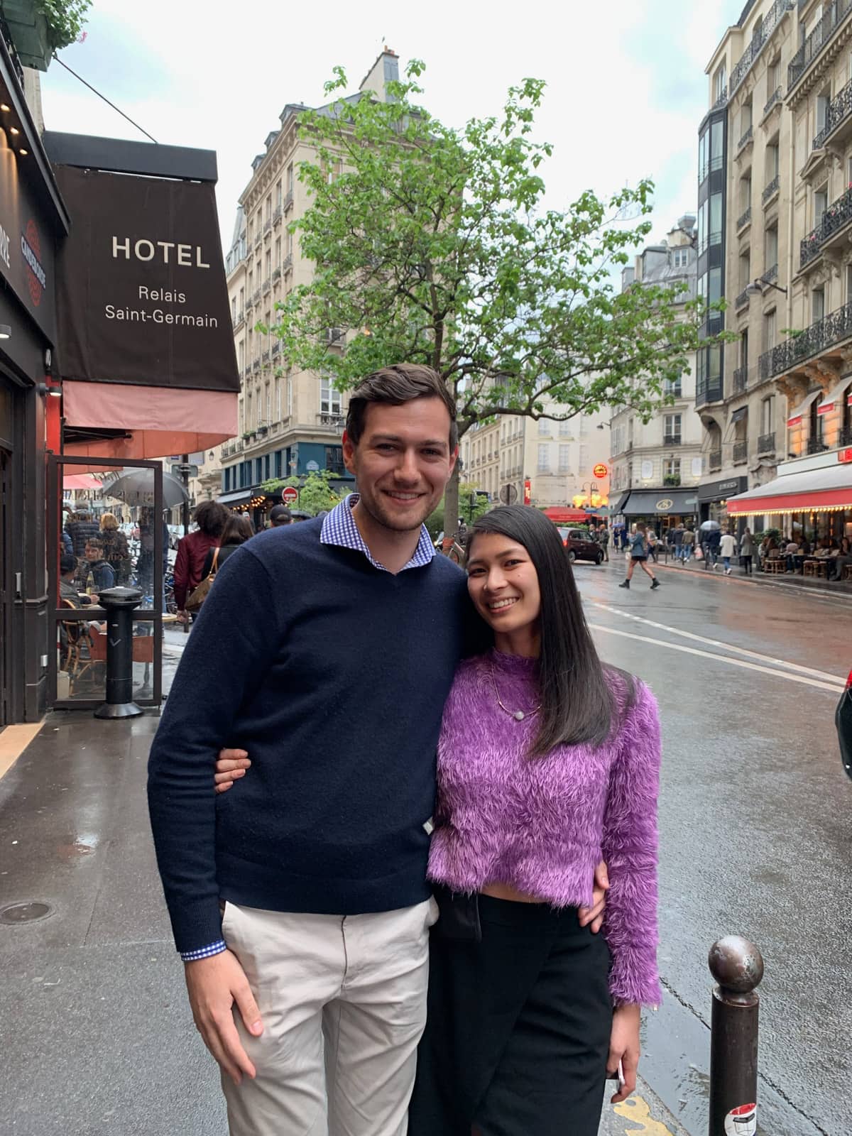 A man and woman with their arms around each other on the sidewalk in Paris. The woman has a purple sweater and black skirt on; the man is in a navy sweater.