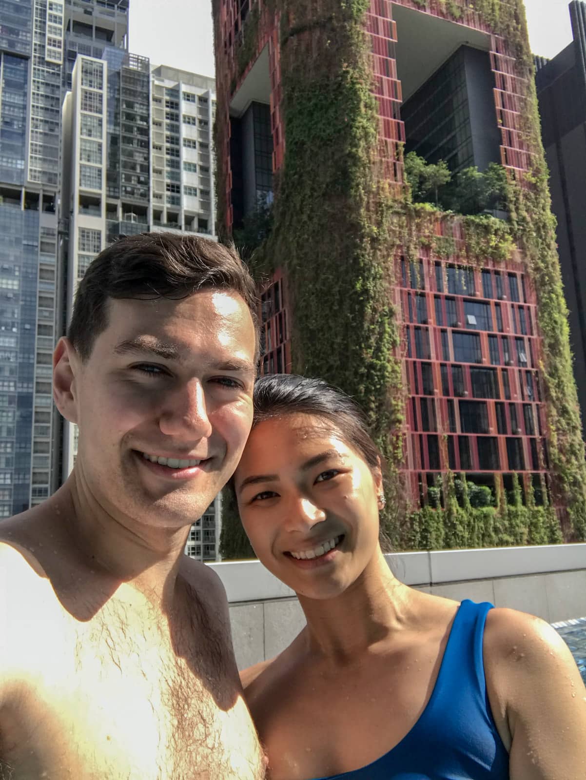 A selfie of a man and woman in a rooftop swimming pool, with a red building behind them that has green plants growing on it. The woman is wearing a bikini top with one strap.