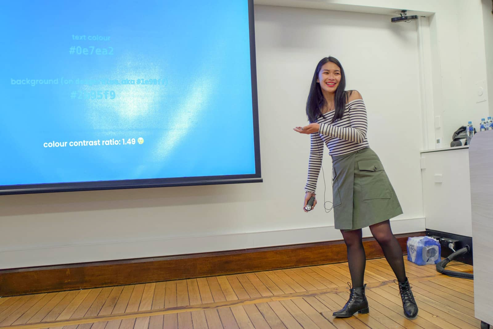 A woman with dark hair, wearing a khaki skirt and striped top, gesturing to a screen with some blue text displayed on it