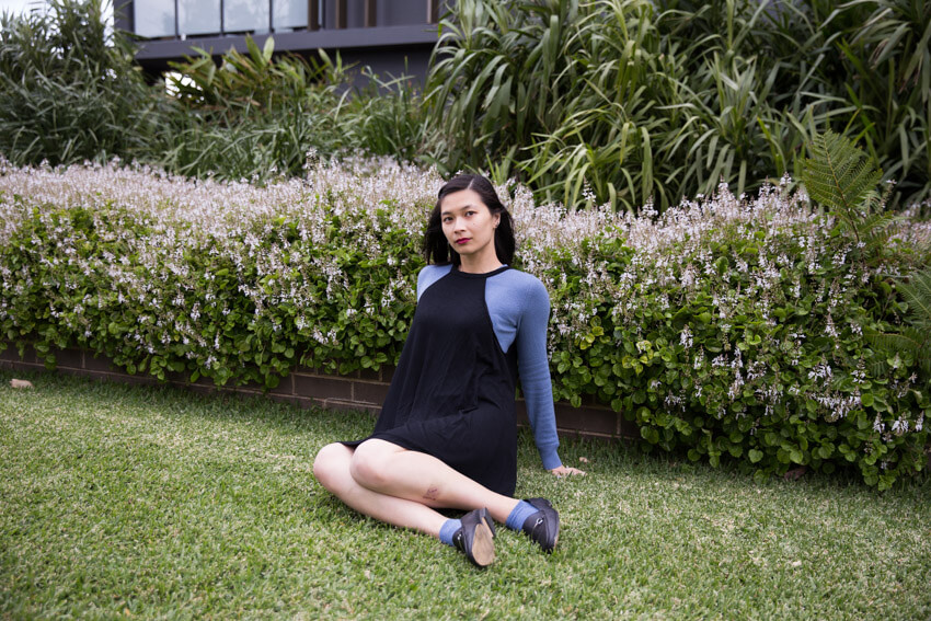 A woman with short dark hair wearing a blue-grey sweater with a black dress over it, sitting on grass with a short brick wall and flowers in the background. The woman has her knees bent to one side, is wearing matching blue socks and black loafers, and is resting with her palms back. She is looking in the distance.