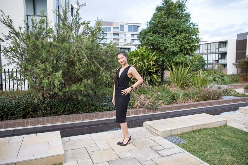 A woman in a black dress with a plunging neckline. She is wearing black pumps (heels) and standing on yellowed concrete. Her left hand is on her hip and her body is angled slightly towards the camera.