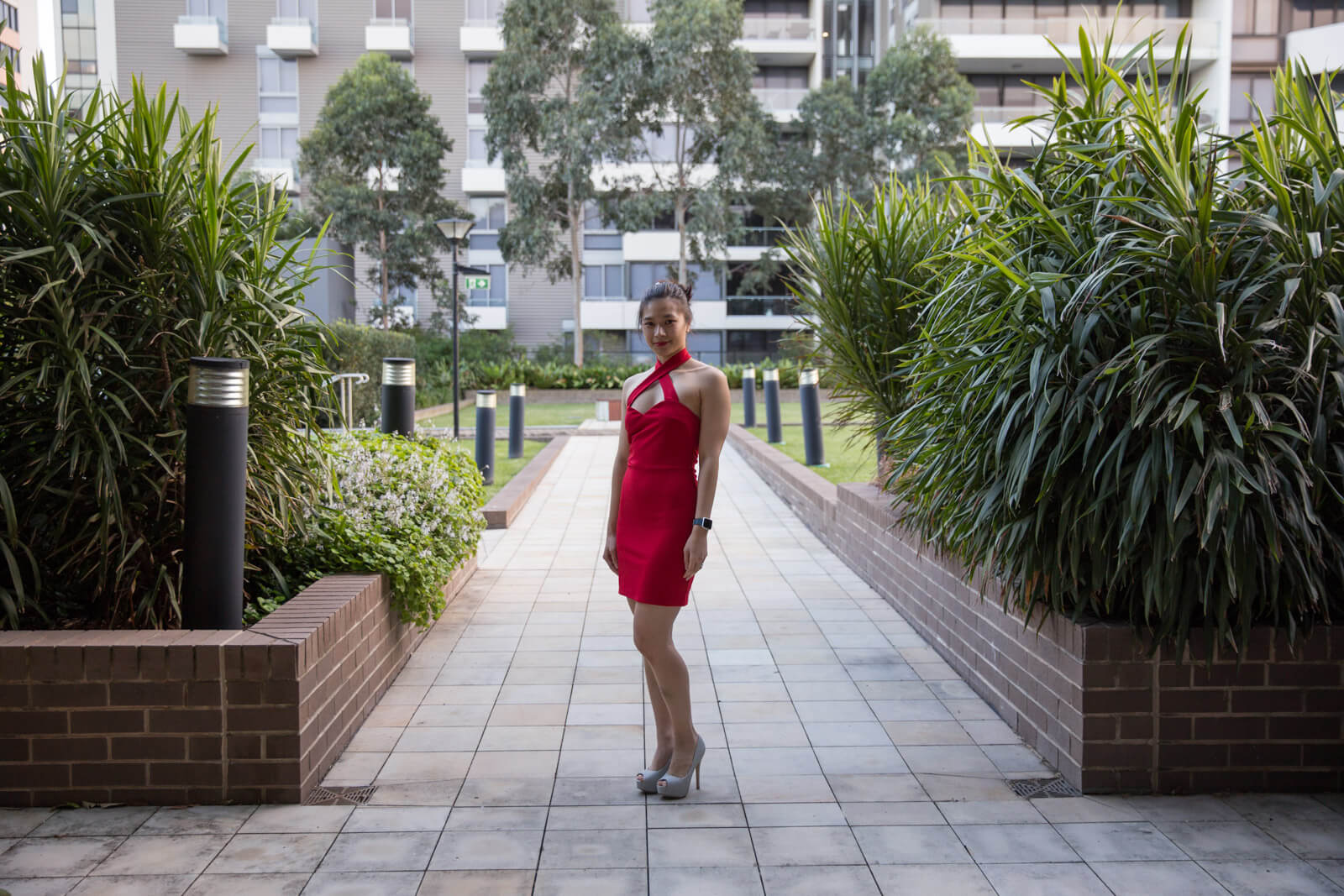 A woman wearing a red dress which has a cross-over front strap above the bust, her dark hair worn in a bun. She is wearing silver high heels and standing on a path with short brick walls and plants and grass outside of the walls.