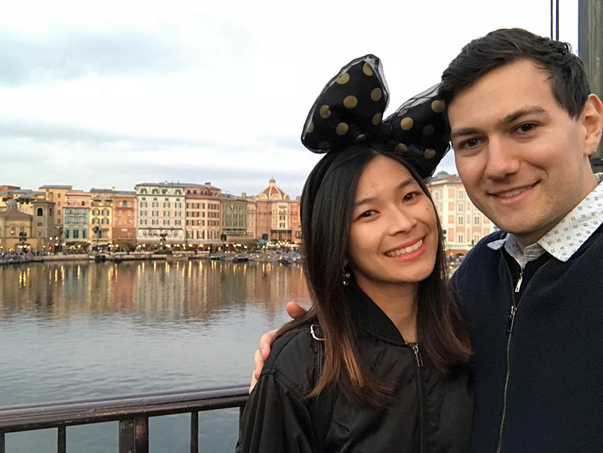 Me and Nick, smiling, with a water port view in the background and old style buildings. I have a headband with a black bow with gold spots, Nick has his arm around me