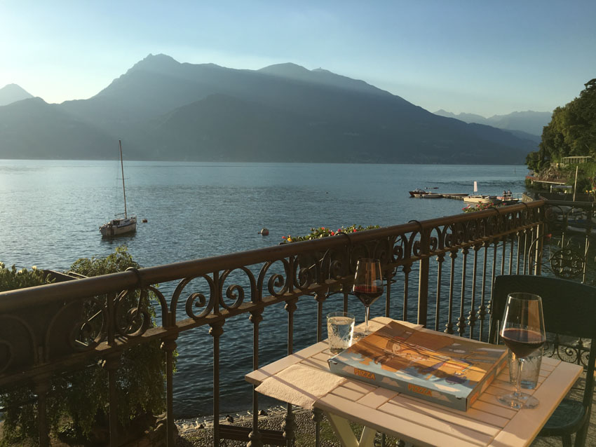 A box of pizza and two glasses of wine on a balcony overlooking a beautiful lake in the afternoon