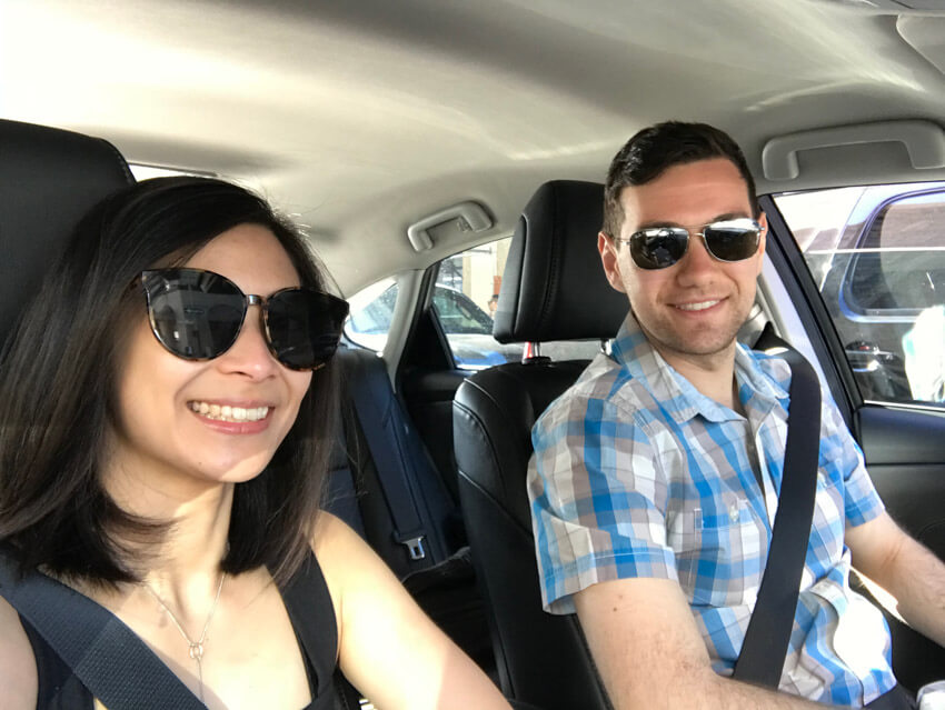 A man and woman with sunglasses, sitting in the front row of a car, smiling