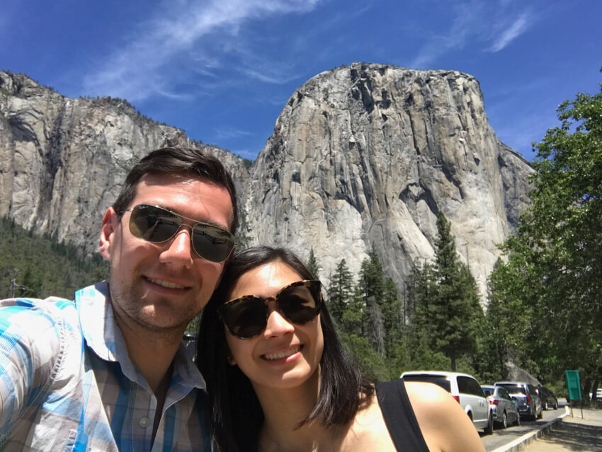 A man and woman wearing sunglasses, smiling, with a large granite rock formation in the background.