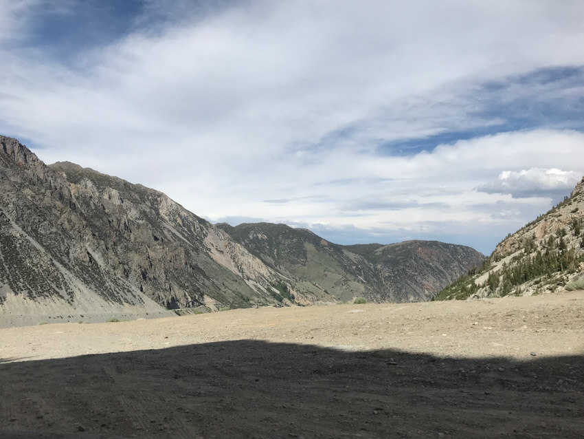 A dusty cliff with deep green mountains in the distance, and the blue sky covered in a huge streak of white cloud.