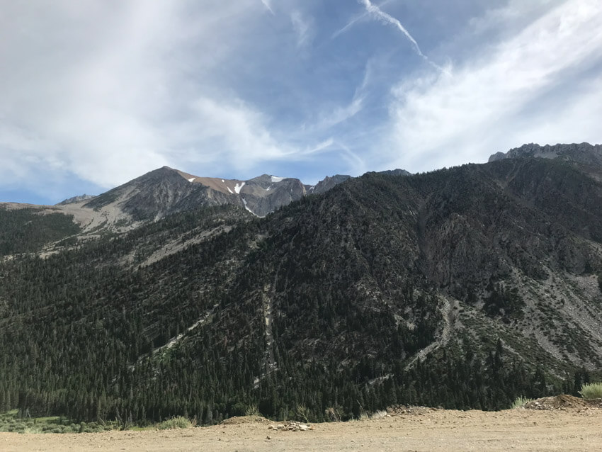 Mountains covered in green trees, with mountains in the background that have patches of snow. The sky is blue but patched with grey clouds.