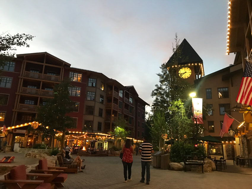 A town plaza at twilight. There is a clock tower and restaurants that are lit with rows of small lights. In the foreground are outdoor chairs, some people sitting and a couple walking.