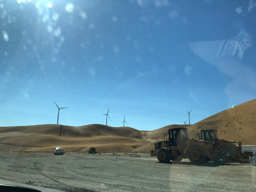 A gravel area with brown hills in the background, topped with windmills. In the foreground are a couple of tractors
