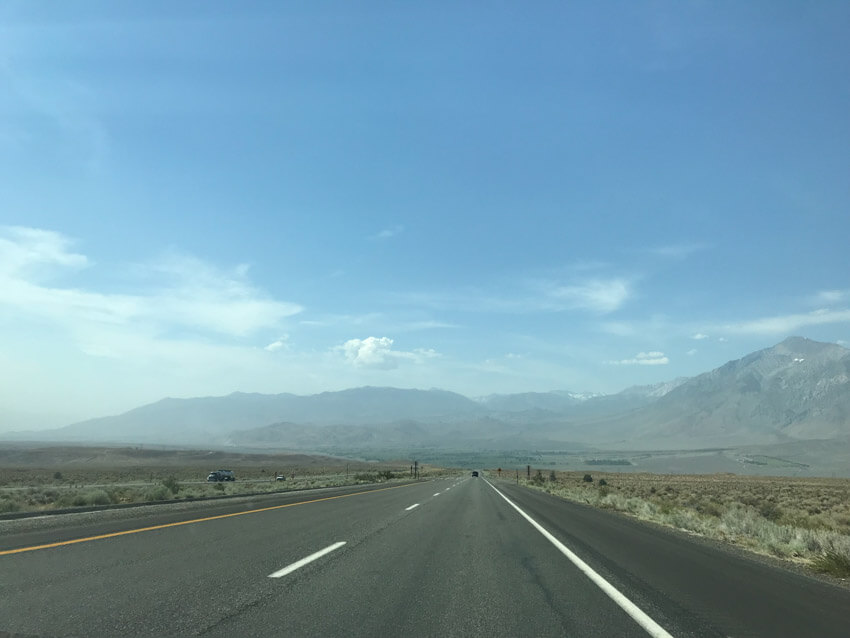 A stretch of road with several cars visible at a far distance. The landscape is hazy with mountains in the backgrounds