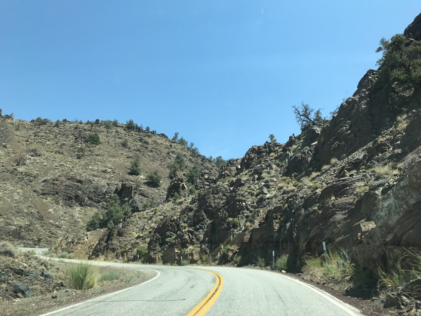 A road that turns towards the left and then right again, in an S-shape. Dark-coloured rock scattered with light green weeds are to the right of the road. In the distance is dirt terrain with small green shrubs.