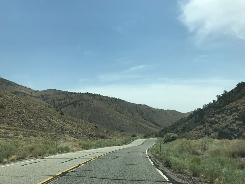 An open stretch of road with light green shrubs to the sides, and dark brown hilly areas in the distance.