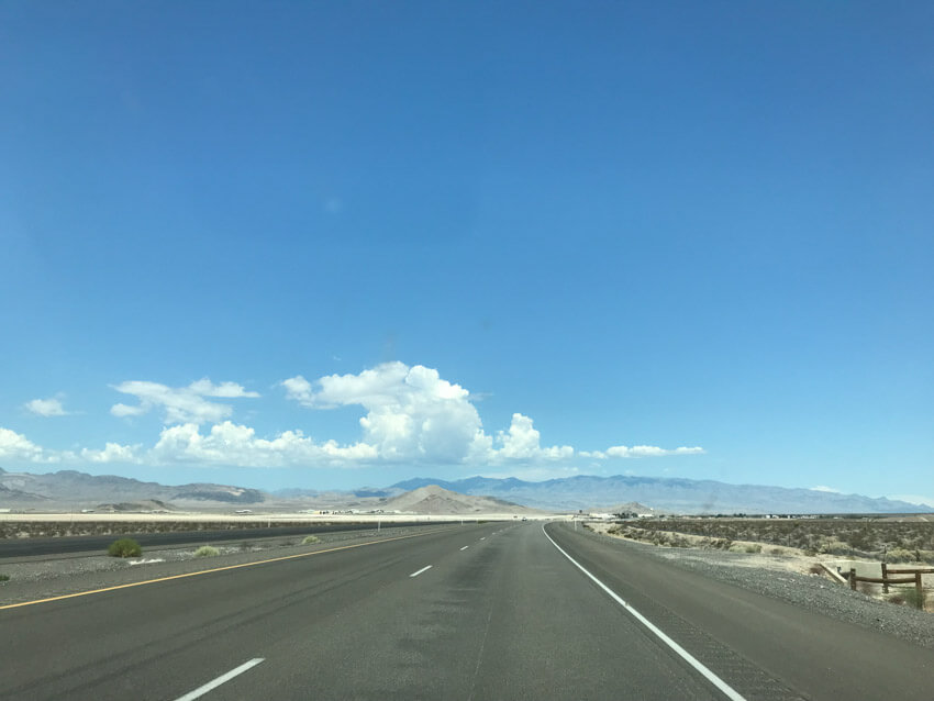 A long stretch of road with desert terrain on the sides. Shallow mountains can be seen in the distance, and a lot of the sky can be seen in the photo. Some white clouds can be seen just above the top of the mountains