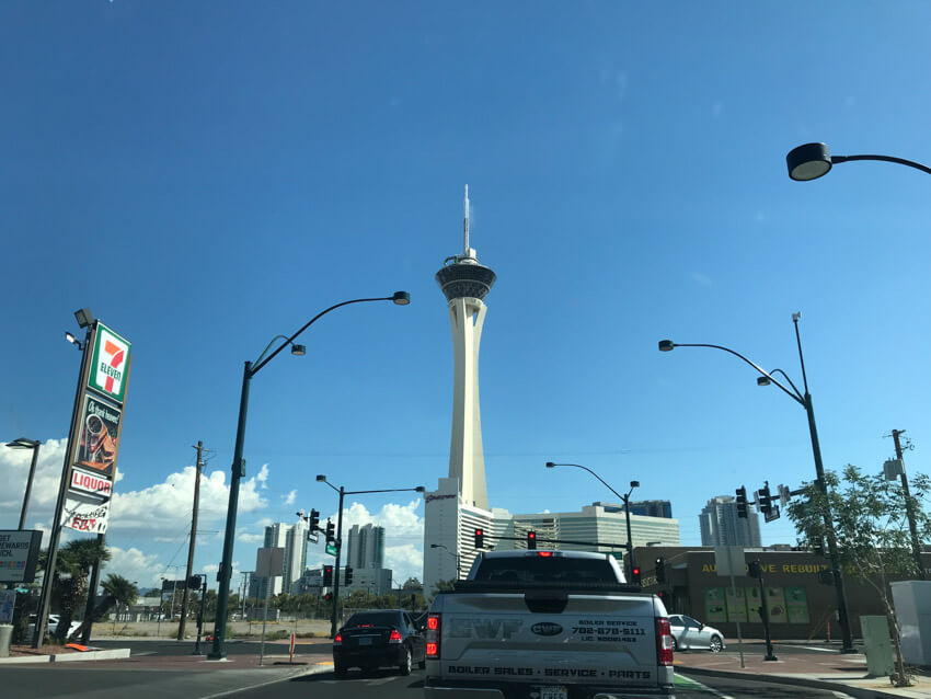 The intersection of a road seen during the daytime, with street lamps on poles visible at the sides of the road. Ahead of the intersection is a tall tower with a pointed top.