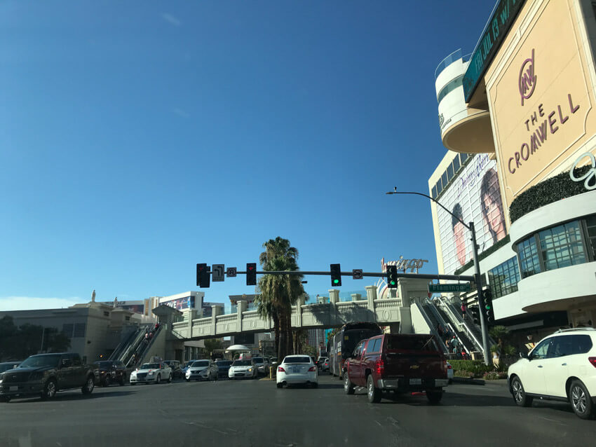 A public road intersection, seen from a car. Cars can be seen travelling in both directions. There is a set of traffic lights hanging from a pole above the height of the cars, and to the right is a large building with “The Cromwell” printed on it