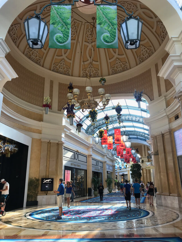 A walkway down a shopping area, with store entrances on the sides. Lanterns hang from the ceiling and there are some people walking down the walkway.