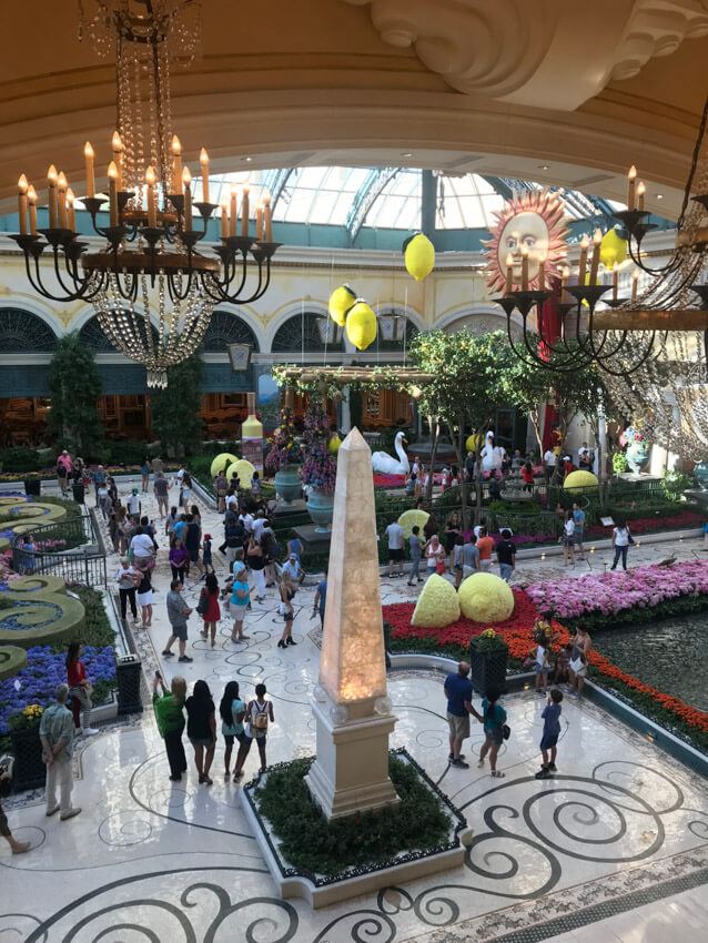 A high view of an area in a hotel with tiled floor. There is a tall pointed stone sculpture in the foreground, man-made gardens and people walking in between the gardens. Some chandeliers and big paper lemons hang from the ceiling.