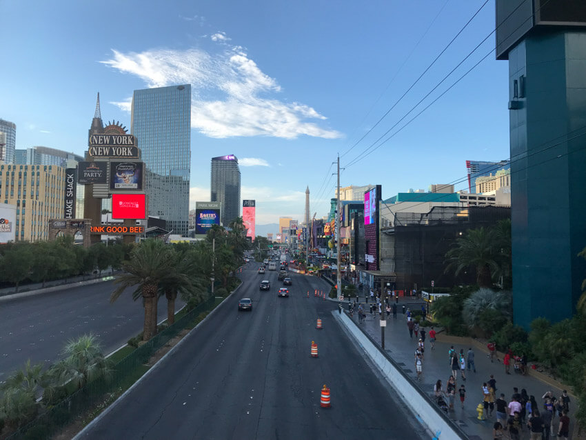 A view of a four-lane road, with modern buildings on the left and right. On the left are lanes with traffic travelling in the opposite direction, and on the right are pedestrians walking on the path