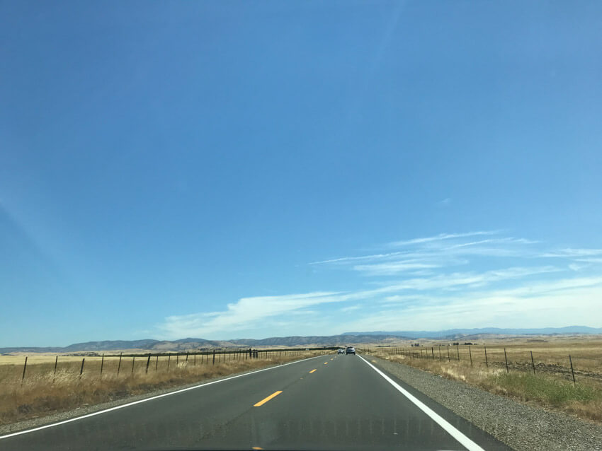 A long stretch of road with blue sky, cars a far way into the distance, and light fencing on the sides.