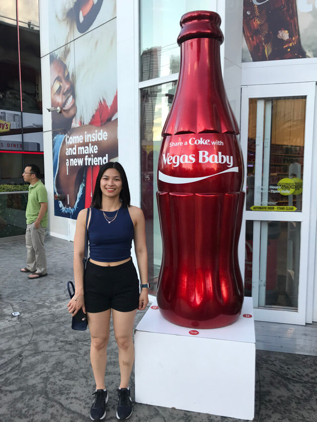 A woman in a blue top and black shorts, standing next to a life-size red Coca Cola bottle reading “Share a Coke with Vegas Baby”