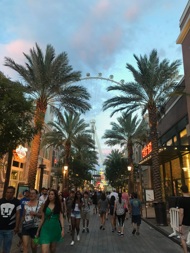 A busy pedestrian street with people walking, palm trees down the sides of the street, and a ferris wheel in the background