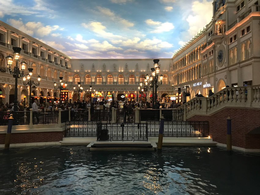 A river with railings, a path, and buildings built up around it, located indoors. The ceiling is painted with blue sky and white clouds to resemble an outdoor setting