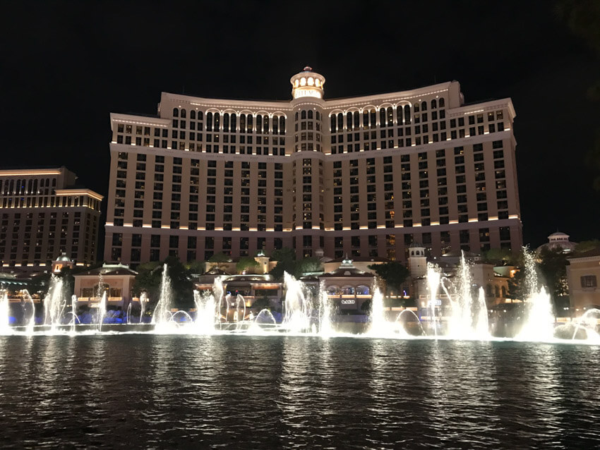 A nighttime photo of a hotel with many windows, with light-up water fountains mid-spray in the foreground