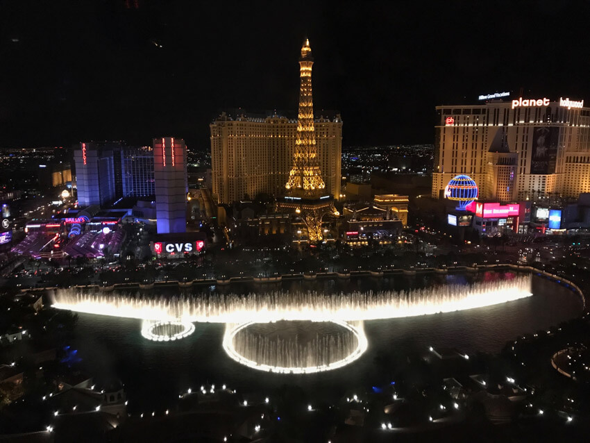 A nighttime view of a city, with an arrangement of lit-up water fountains. In the background is a replica of the Eiffel Tower